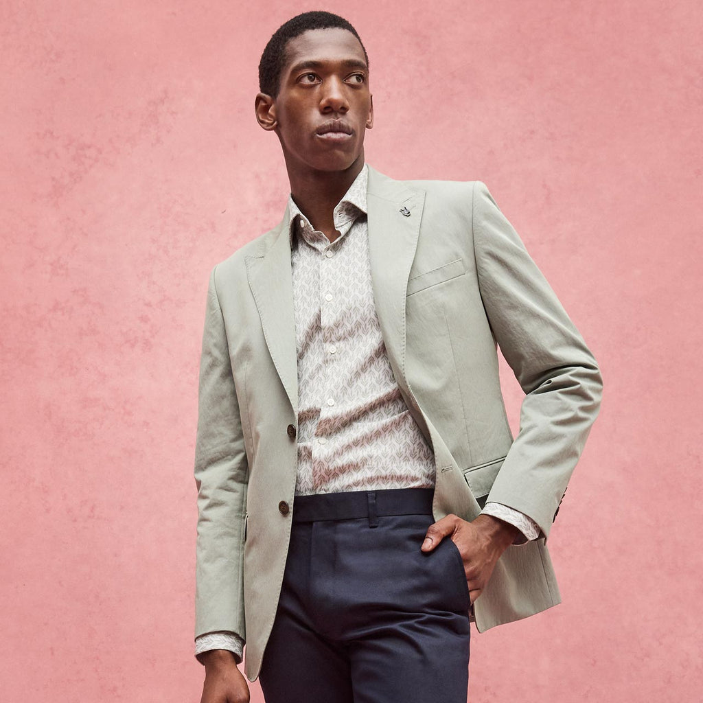 Man wearing a light gray blazer over a patterned shirt with navy pants against a pink background